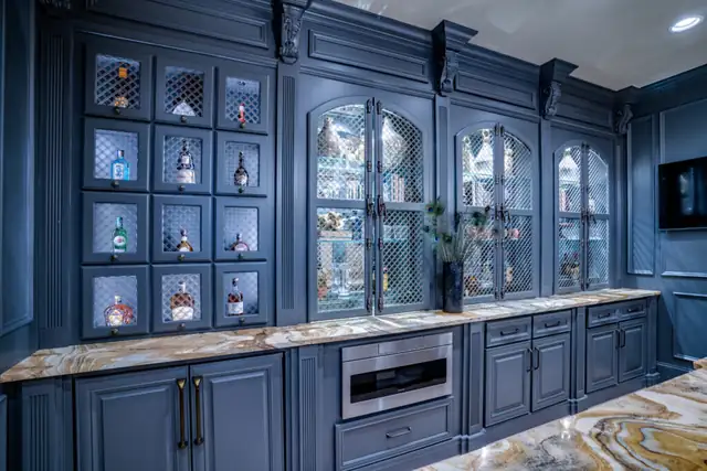 Navy blue refinished cabinetry with lit glass display cubbies, arched glass doors, decorative crown molding and a dramatic stone counter
