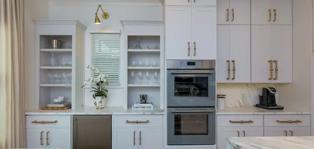 White refinished kitchen cabinets with gold hardware, open glass-front uppers, a brass sconce and double wall ovens