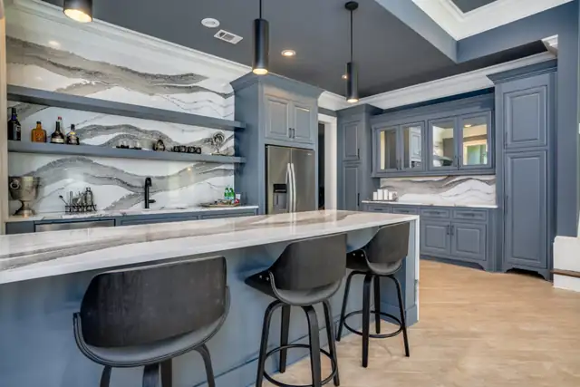 Basement bar with navy cabinets, a marble waterfall counter and backsplash, floating shelves and black counter stools