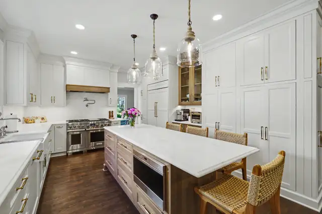 White kitchen with a long marble-topped island, woven stools, tall pantry cabinets, a wood range hood and glass pendants