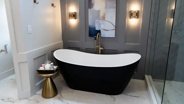 Remodeled bathroom with a black and white freestanding tub, gray paneled accent wall, brass sconces and marble-look floor tile